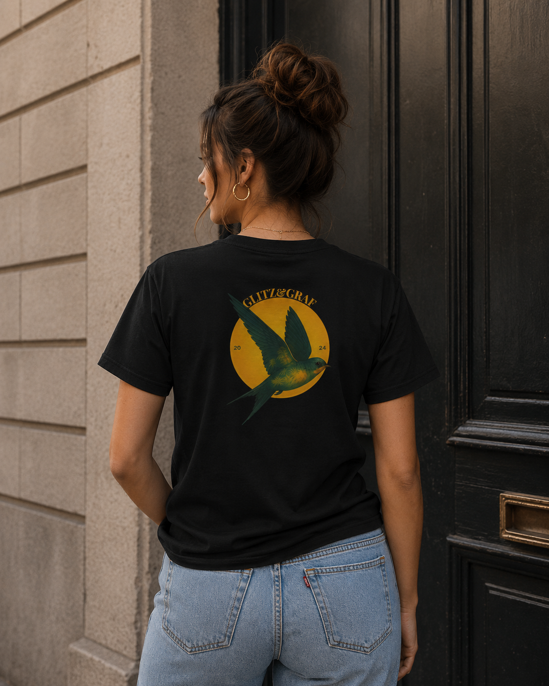 Person wearing a black t-shirt with a graphic design on the back, standing against a building.