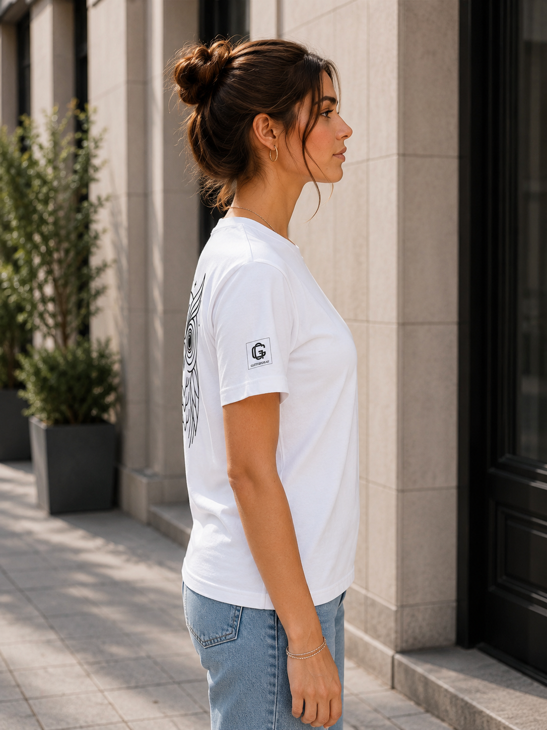 Woman wearing a white t-shirt with a logo on the right sleeve and a owl graphic on the back, standing outdoors.